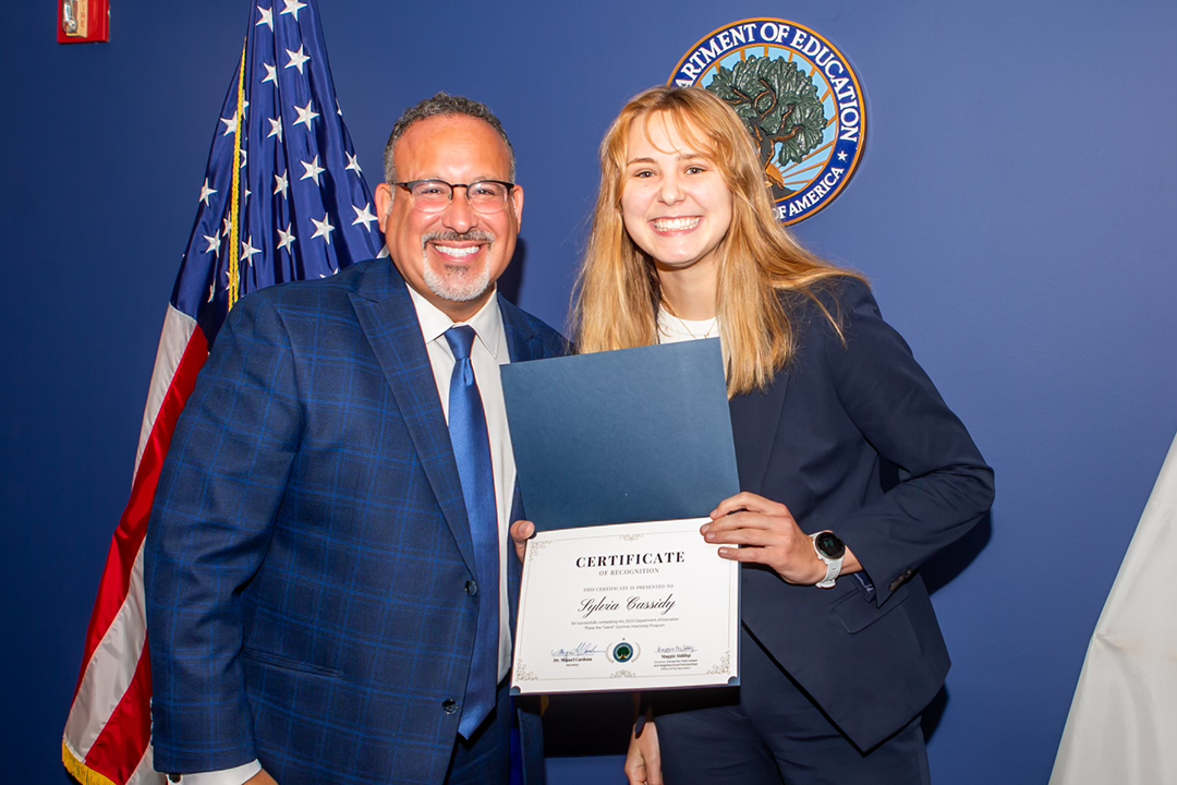 Sylvia Cassidy smiling and holding up a certificate next to a man in a suit with a Department of Education seal in background